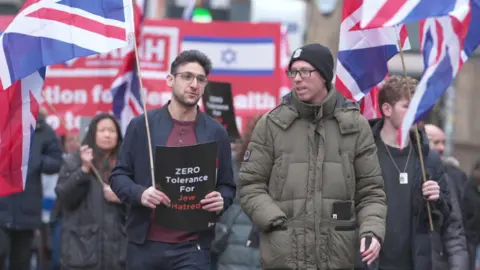 Yoni Finlay, who is holding a Union Jack flag and a sign which reads "Zero tolerance for Jew hatred", is walking alongside other protesters at the march. 