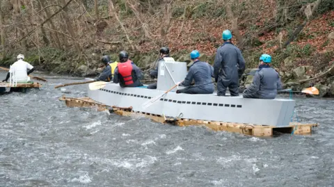 Sam Hepburn A raft shaped like a grey submarine with six people sat on top of it holding oars.