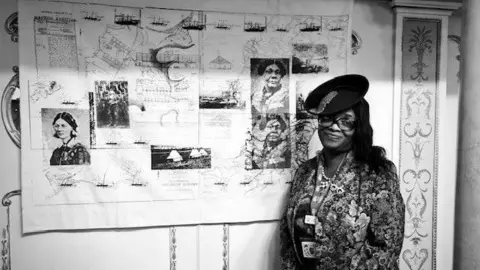 Jacqueline Braithwaite A black and white image of Jacqueline Braithwaite, wearing a black hat, glasses and a floral dress, standing in front of her artwork. The faces of Florence Nightingale and Mary Seacole can be seen.