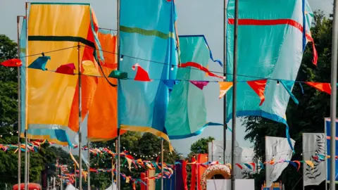 Bridge yellow and blue festival flags with ribbons in similar colours fluttering underneath.  The festival is based among green trees.