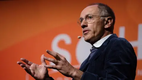 Getty Images Michael Moritz is on stage, speaking with his hands in front of him with an orange background behind him. He has a microphone headset by his mouth and is wearing a navy corduroy jacket with a blue jumper and white shirt underneath.