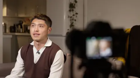 Andy Hu wears a white shirt and brown tank top. He is looking off to his right while a photographer frames him on a camera prior to filming. 