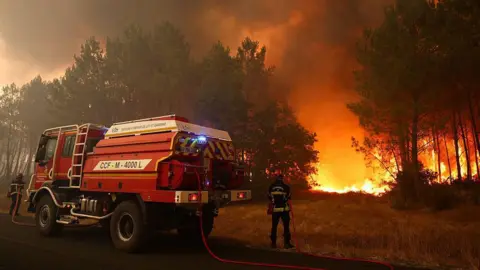 Firefighters try to extinguishing fires burning trees using a wildland water truck. The flames can be seen in the forested area