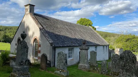 A small church building with white washed walls and a slate roof. There are many old grave stones covered in mosses and lichens. 