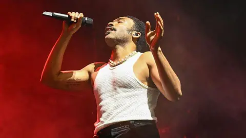 Donald Glover, wearing a white tank top and holding a microphone on stage with smoky red background