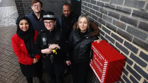 Oxford City Council Councillor Lubna Arshad (first from left on the first row), a TVP officer, writer and performer Claire Nelson, and two men behind them smiling for the camera next to a bleed kit.