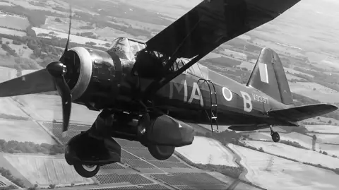 A black and white image of a World War Two plane mid-flight. It is banking slightly to its right. It has a propeller on its nose and has various RAF letters and numbers painted on its body. A patchwork of fields is on the ground.