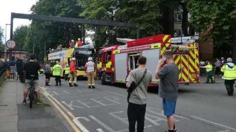 Tyler Tyldesley People stand around looking at a fire truck which is parked in front of a yellow double decker bus which has had its top ripped off and the top windscreen smashed. Firefighters in helmets and high vis are standing on the exposed top deck.