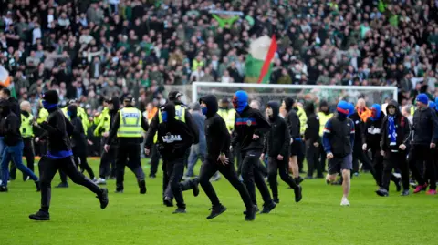 PA Media A number of football fans running across a football pitch wearing black clothing and face coverings.