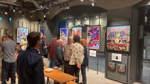 A group of people standing in a room that is full of Ipswich Town posters, in frames around the room.