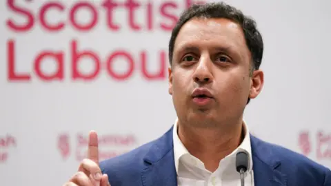 Anas Sarwar, with dark hair and wearing a blue suit and white shirt, speaks at a Scottish Labour press conference with his right index finger raised 