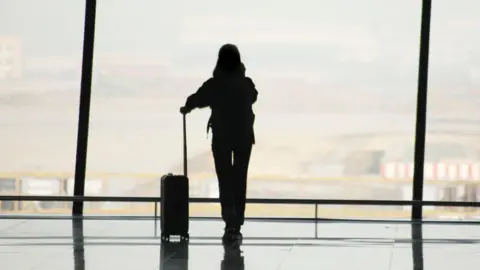 Getty Images The silhouette of a person with a suitcase standing at a big glass window at an airport 