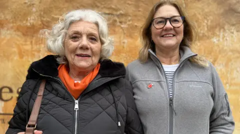 Emma Baugh/BBC Jackie, a women with shoulder length light brown hair, wearing a grey jacket standing next to her mum, a woman with short white hair, wearing black jacket and an orange scarf.