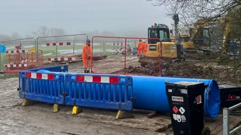 Workers in orange uniforms and helmets stand beside excavators behind barriers in a misty field