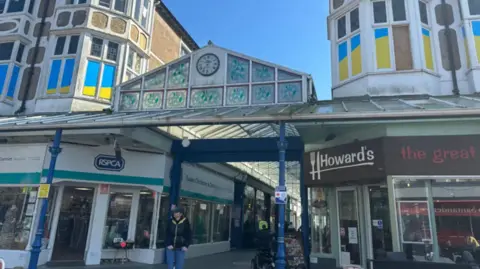 Sophia Cobby/BBC The entrance to the glass roofed arcade which has shops on both sides. It has an old clock with Roman numerals and turquoise stain glass panels