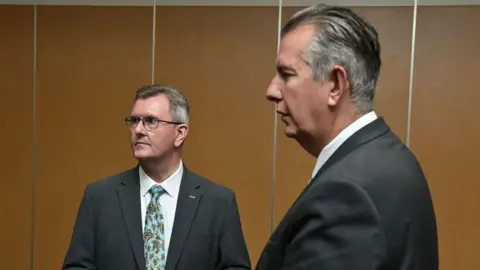 Getty Images Two men look off to their right. In the foreground is a tall man with slicked back grey and white hair. He is wearing a dark suit and white shirt. The shorter man in the background can be seen more visibly. He is wearing a grey suit, white shirt and turquoise patterned tie. He is also wearing glasses and has a fish badge pinned to his jacket. They are both standing in front of a brown wall.