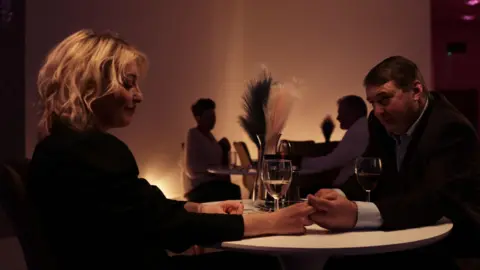Two people sat over a table in a restaurant. They are holding hands. There is a glass of wine in the centre of the table.