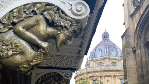 A wood carving of pan, with pipes, is painted gold and sits under a porch - in the background the domed top of the Radcliffe Camra can be seen.