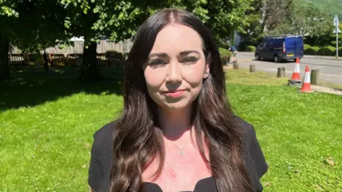 A woman with long black hair and a black dress stand outside on a grassy area