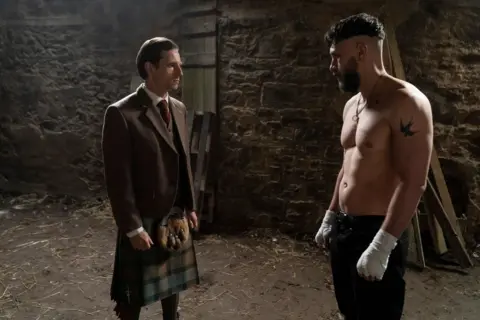 BBC/Mam Tor Productions/Anne Binckebanck In a barn a man in a suit jacket and a kilt stands opposite a man wearing jeans with his shirt off and bandages on his hands.