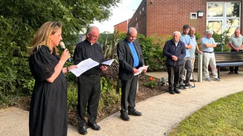 A number of people taking part in reading at a service at the memorial garden. A woman in black is leading the readers and two ministers are holding books waiting to read from them. Members of the public are watching and listening in the garden.