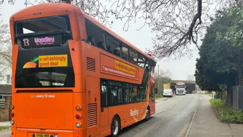 A double-decker bus is pictured from behind. The upper deck of the vehicle has sustained significant damage and is tilted backwards. Many of the windows appear to be smashed. An electronic display reads "13 Woodley". A poster below the display reads: "a refreshingly good deal". 