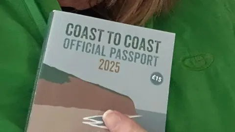 Wendi Merrit A hand holding a small booklet title 'Coast to Coast official passport 2025' against a green background