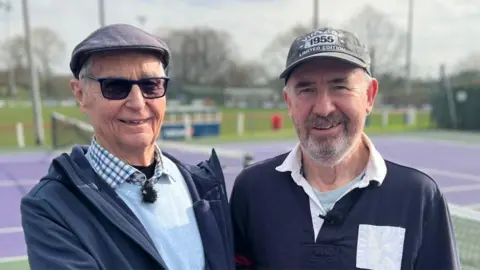 Head and shoulders image of David Pearson and Andrew Lockwood standing outside at Garstang Tennis Club by a purple court. David is wearing sunglasses, a flat cap, and a blue checked shirt under a pale blue jumper and dark blue coat. Andrew is wearing a baseball hat and navy and white rugby shirt. He has a short greying beard