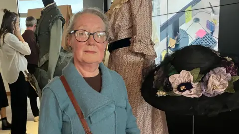 Julia Fox wearing a blue coat and looking at the camera. To her right is a black hat on a stand. Behind her is a dress.