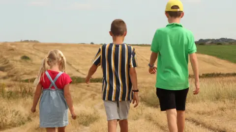 Three children, a little girl, a boy and an older boy, walk along dry grass near the coast. They have their backs to the camera. It's a summer's day.