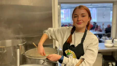 Mark Ansell/BBC A young woman wearing an apron uses a wooden spoon to stir some food in a pot.