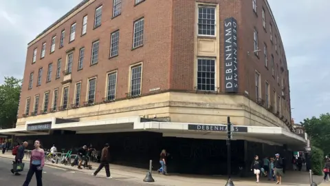 Owen Sennitt/BBC A large department store building can be seen, with people walking past. The ground floor entrances have been boarded up by black hoardings covered in graffiti.