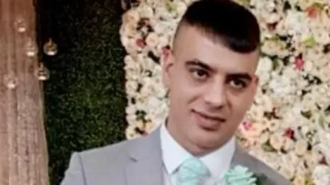 West Yorkshire Police Choudry dressed in formal wedding attire, standing indoors against an elegant floral and greenery backdrop. He is wearing a light grey suit paired with a white dress shirt and a patterned white waistcoat. A mint‑green tie and a matching boutonnière add coordinated colour accents to the outfit. The background includes decorative drapery, floral arrangements.