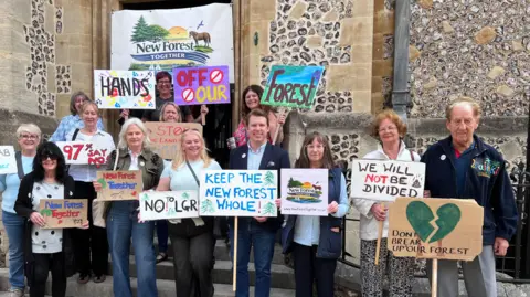 Group of people stand outside Hampshire council chamber with signs saying "don't break up our forest", "Keep the New Forest whole" and "we will not be divided"
