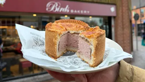 BBC A large pork pie with a quarter cut out sits on a wrapper and plate in front of a bakery. The plate is being held be someone out of shot. The bakery has a large glass front and the maroon sign reads 'Birds Bakery.' There is a pavement and road sign beside it.