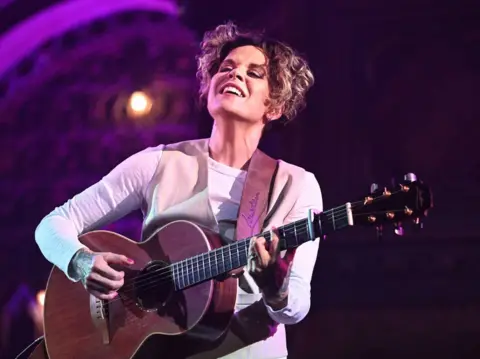 Getty Images Amy Wadge strums a guitar against a purple backdrop