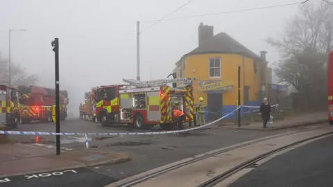 Fire trucks next to a yellow building. There is blue police tape stretched across the highway intersection.