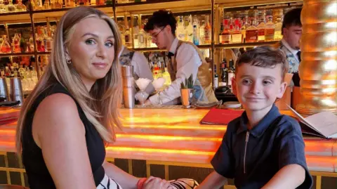 Zoe Stokes-Jones Photograph of Zoe Stokes-Jones and her son, Jacob, pictured sat at a bar of a restaurant. They are both sat on stools and turn around to look at the camera. Both are smiling, Zoe has blonde long hair parted in the middle and blue eyes. Jacob wears a navy polo shirt and smiles at the camera. He has brown short hair.