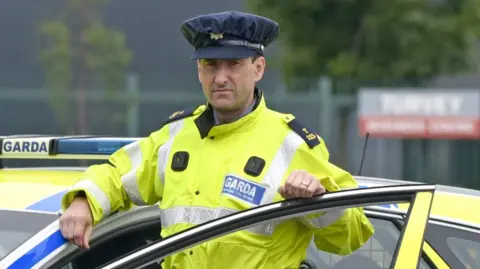 An Garda Síochána A man wearing an Irish police officer uniform of a yellow hi-vis jacket standing behind the open door of a police car which is stationary.