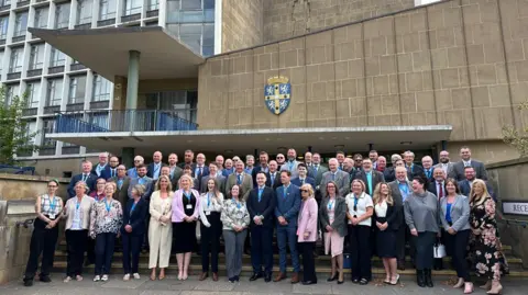 The new council members smiling as they line the steps in front of Durham County Hall. It is a beige building with a lot of windows and the county crest.