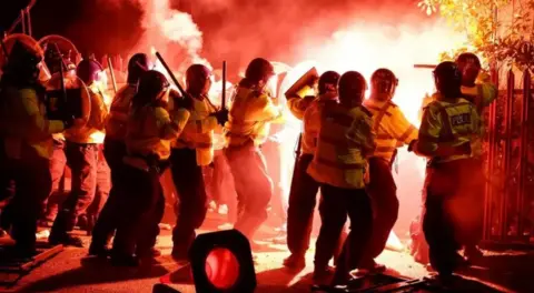 Police officers in riot gear with shields with a red flare going off in the background