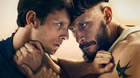 Jamie Bell and Richard Gadd in character as Niall and Ruben in Half Man. Their foreheads are touching, their facial expressions are stern and their arms are raised to each other's necks, with their fists clenched.