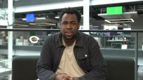 A man in a dark denim jacket and a beige t shirt is sat down looking straight at the camera. He has spiky afro hair and there is a small mic poking out of his t-shirt. In the background there are blurry tv screens with various different output from the BBC. There is also a glass wall roughly a waist height and two coloured squares, one blue and one green.