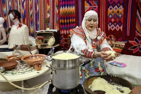 APP/NurPhoto via Getty Images A woman, wearing a colourful outfit, scoops up couscous