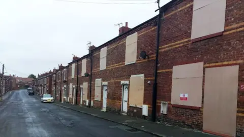 A row of red-brick houses with almost every window and door boarded up