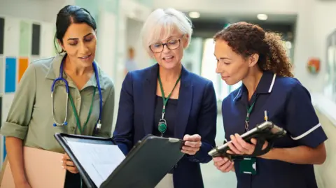 A woman middle-aged woman wearing a shirt with dark hair and a stethoscope around her neck, an older woman in a blazer holding a file, and a younger woman in a nurse's outfit holding a tablet computer, all stand next to each other in a hospital ward