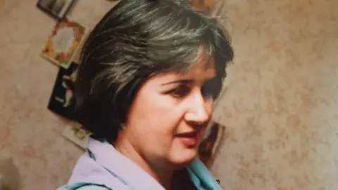 Family photo Archive side profile photograph of Judith Rhead. She has brown short hair, fringed at the front. Behind her a wall covered in cards can be seen. She has brown eyes. 