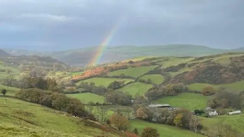 NFFN A rainbow is pictured against grey clouds over green rolling hills