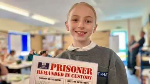 BBC/ Nicola Rees A young white girl with blonde hair tied back in a pony tail is wearing a grey prison jumper. She holds a card up with the words 'prisoner remanded in custody' on it and smiles.