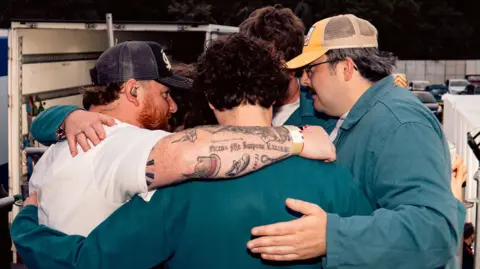 Hope Simmers Cammy Barnes, wearing a black cap and a white T-shirt, has arms around band members and crew behind the main stage at Belladrum.